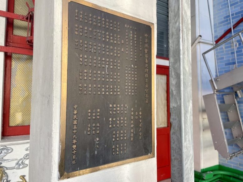 A bronze plaque with Chinese characters mounted on a white stone pillar near a red door and metal staircase.