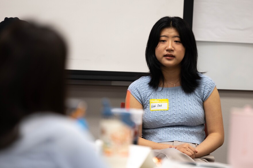 A student is sitting at the front of a classroom facing teachers. She is wearing a light blue shirt and a name tag. 