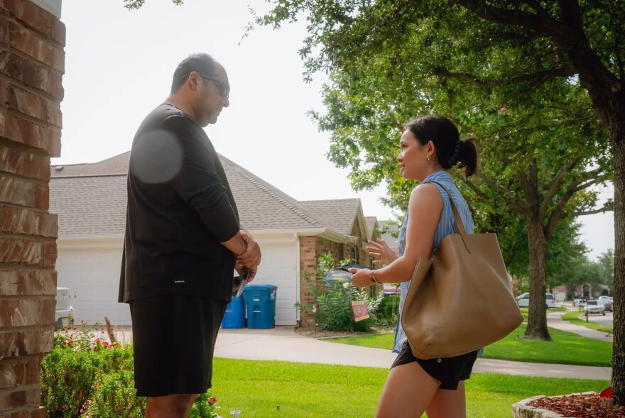 A man on the left stands outside a home while he listens to a woman speaking on the right.