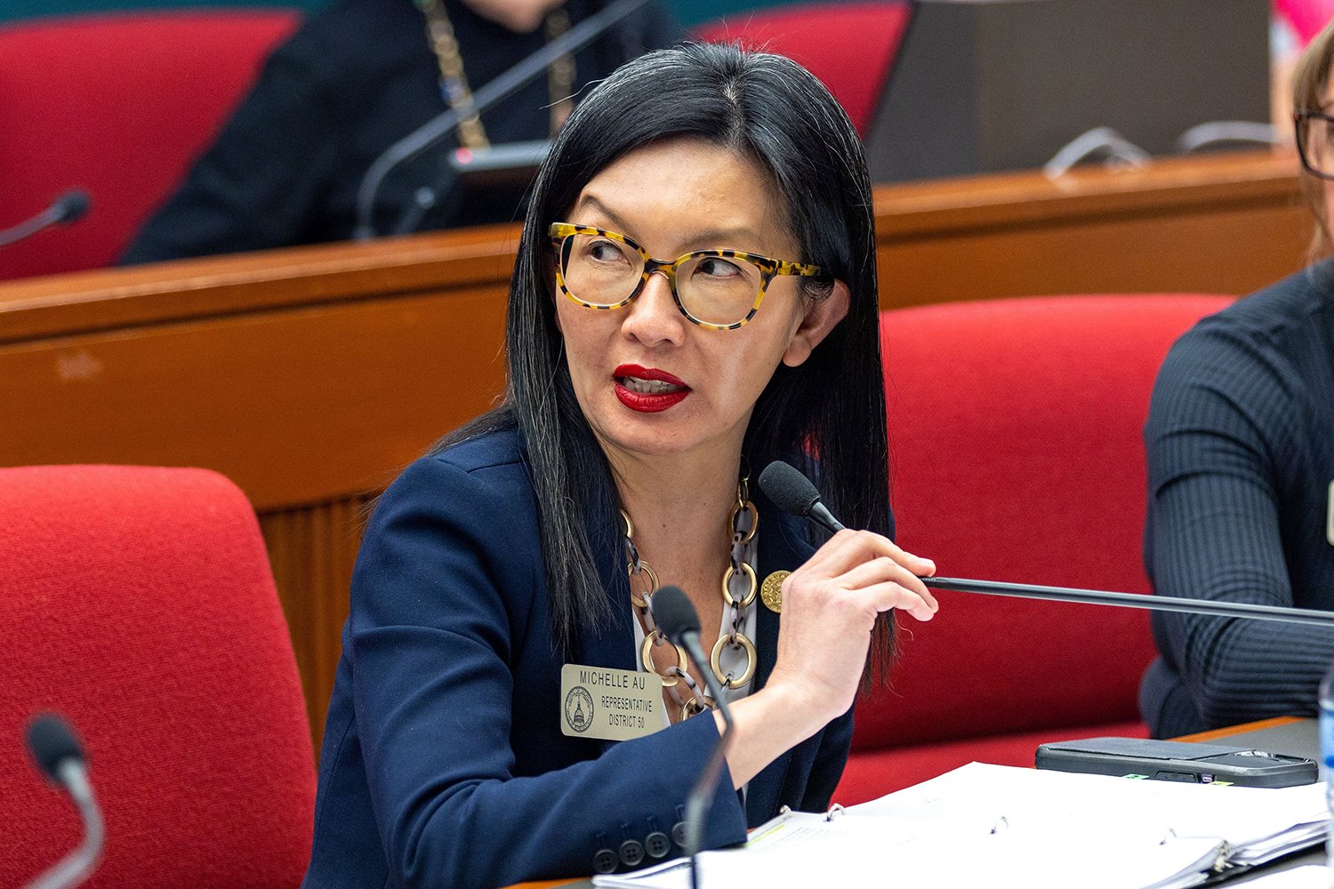 Michelle Au wearing glasses and a suit jacket adjusts a small microphone as she talks in a legislative chamber.