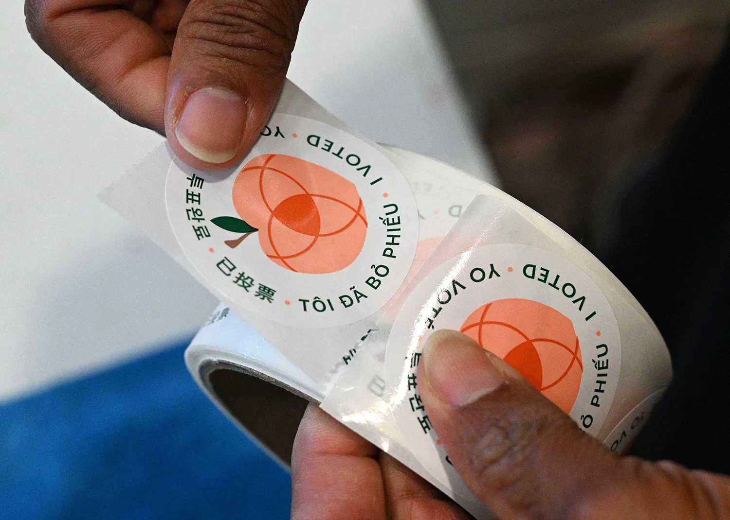 A closeup view of a roll of I Voted stickers with a peach logo on them and messages in multiple languages.