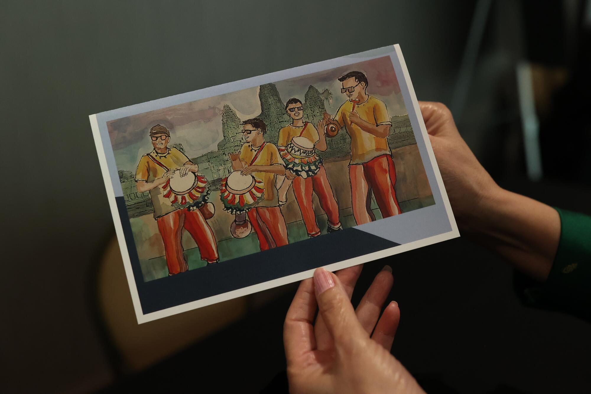 Sithary Ly holds a post card showing man dressed in traditional Cambodian clothing playing on skor thom drums.