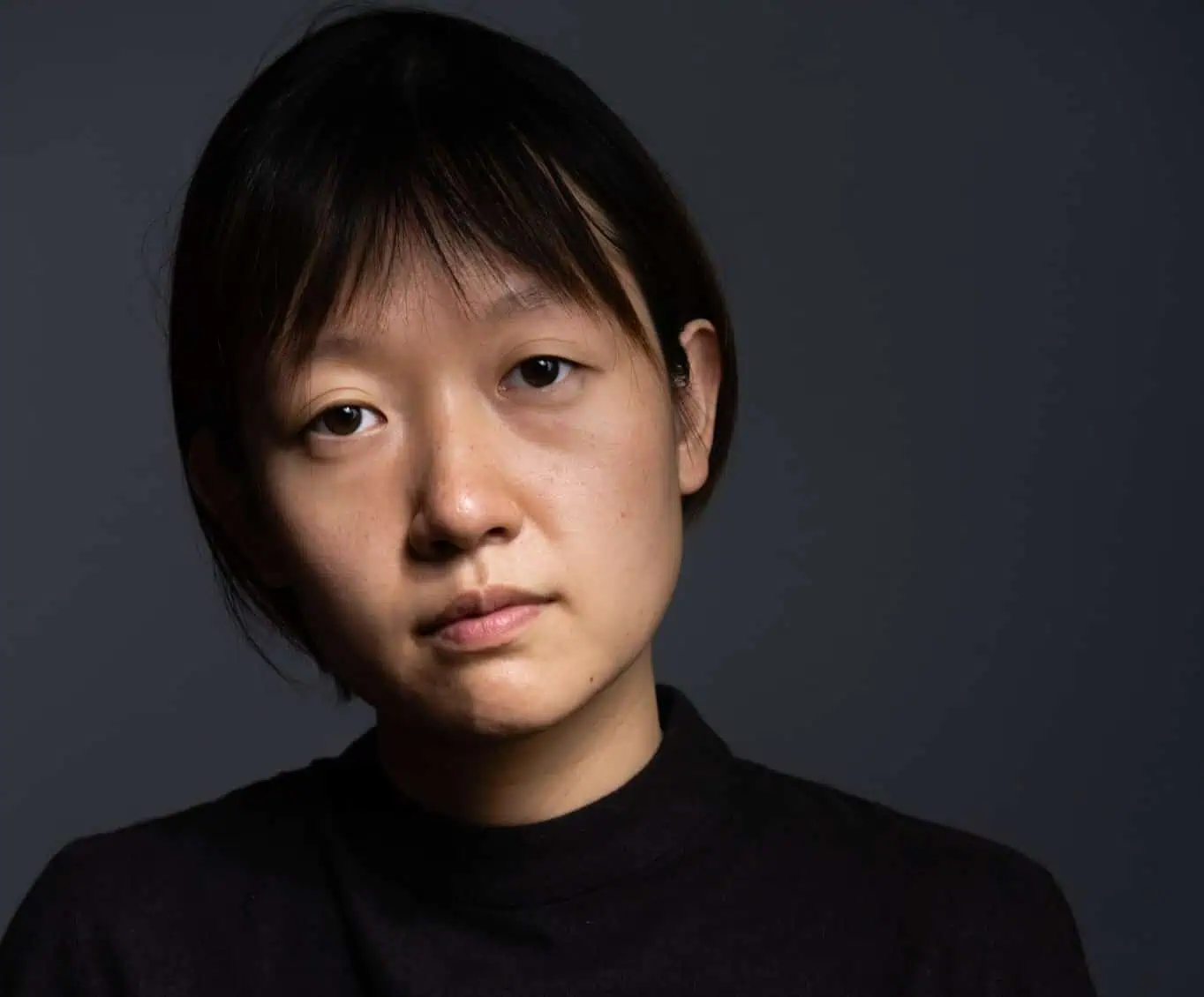 An Asian woman with short hair, in a black top, against a dark gray backdrop.