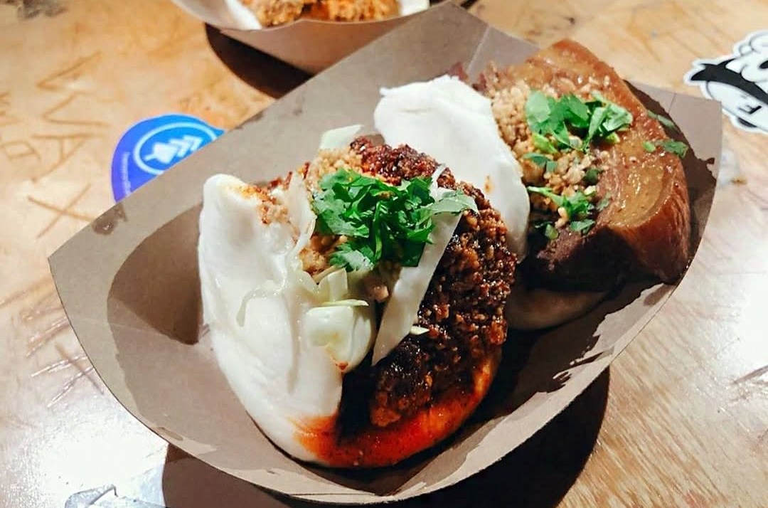 Baos stuffed with spicy fried chicken and pork belly, in a paper boat on a wooden table tagged with various stickers and knife carvings.